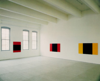 A portrait oriented rectangular red canvas with a black horizonal strip slightly below middle hangs between two windows on th left wall. On the adjacent white wall to the right, two rectangular canvases are hung apart from each other, one red, the other yellow, each with a vertical black wide stripe in the middle.