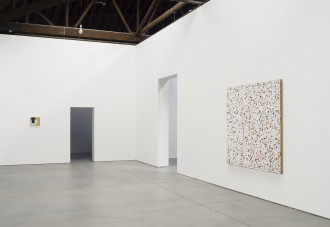A very small work of yellow, black, and white hangs on a far white wall and a very large work of yellow, red, and white hangs on a near white wall with two doorways positioned between them.