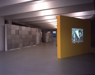 Four large rectangular sculptures stand in an industrial space. Three are decorated with gray 3D geometric patterns and the fourth, painted yellow, features a projected black-and-white image of two people speaking in front of a building.