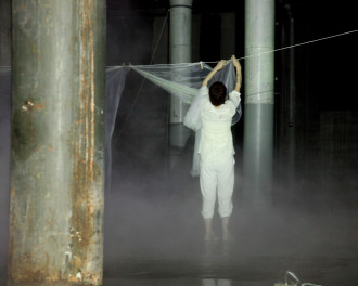 A shadowy image of a person dressed in white in an industrial space with pillars and white smoke near the ground, reaching up to hang sheets of plastic on a string with clothespins.