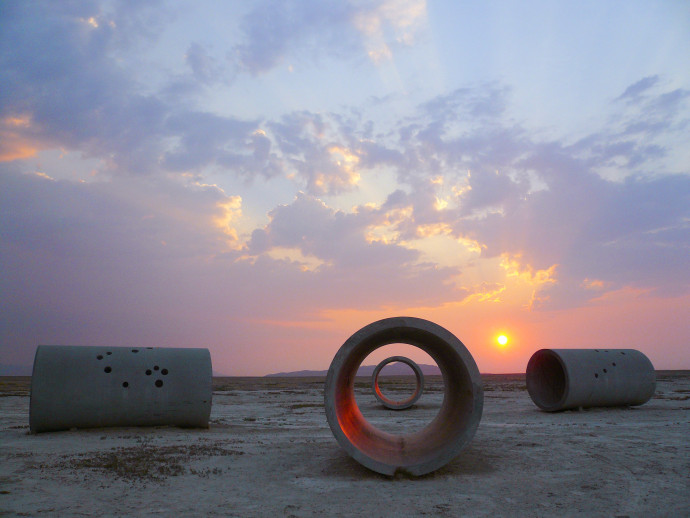 Four large concrete cylinders lie on their sides in the desert. The sun is casting a pink glow along the horizon of an otherwise blue sky.