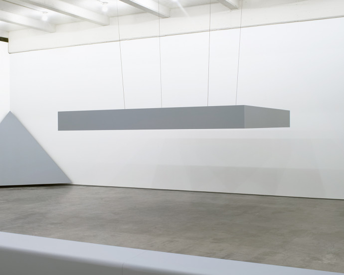 A horizontally oriented, tabletop-like, gray sculpture is suspended from the ceiling.