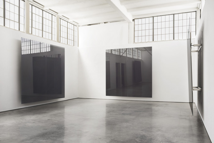 Three large reflective planes of gray sheet glass each mounted to a different white wall in large sunlit room with a polished concrete floor and windows.