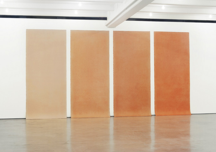 Four long paper works installed in a row on a white wall, the bottoms dragging slightly on the concrete floor. They are all in tan and orange shades, arranged from lightest to darkest staring on the left.