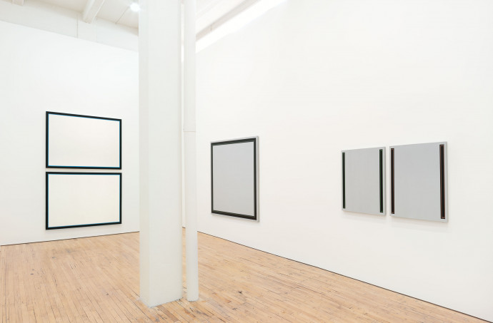 A large room with white walls and a wood floor. To the left of a white column two hortizonal white canvases with black borders are stacked. To the right on the adjacent wall is a large gray canvas with a black border, and two square canvases with black lines on each outer vertical edge.